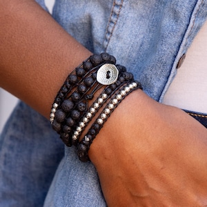 May include: A multi-layered bracelet with black lava rock beads, silver beads, and a silver button charm. The bracelet is worn on a person's wrist.