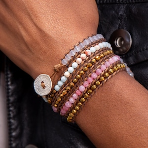 May include: A multi-layered bracelet with pink, blue, and gold beads, wrapped around a brown leather cord. The bracelet has a silver button closure.