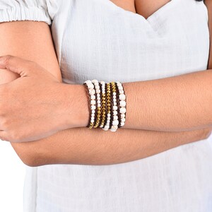 May include: A stack of bracelets with white beads and gold accents on a brown leather base.