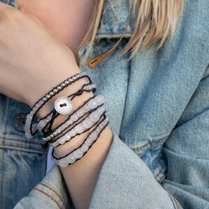 K&ouml;nnte beinhalten: Ein Satz &uuml;bereinander gelegter Armb&auml;nder mit wei&szlig;en Edelsteinperlen, schwarzem Band und einem silbernen Knopf-Anh&auml;nger.