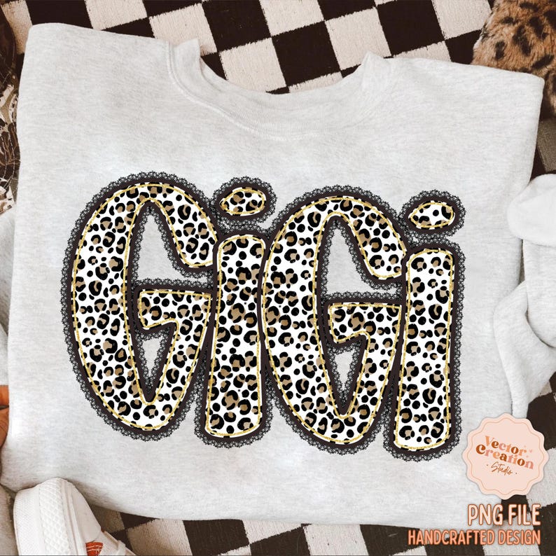 May include: A light gray sweatshirt featuring the name "Gigi" in a bold, leopard print design. The letters are outlined in gold and framed with black lace. The design includes a small, pink, round logo with text.