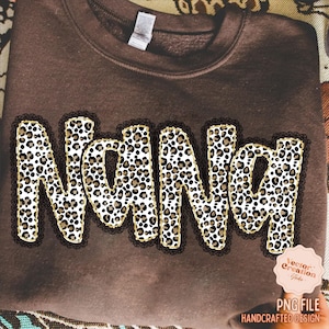 May include: A brown sweatshirt with the word "Nana" in a leopard print design. The letters are outlined in gold and framed with a black lace trim. The bottom right corner features a logo that says "PNG File Handcrafted Design".