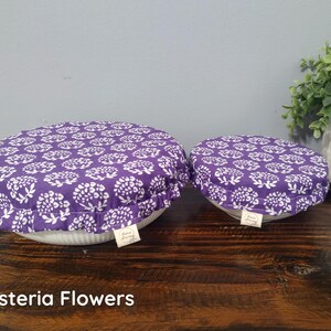 May include: Two round fabric bowl covers with an elasticized ruffled edge, featuring a purple and white floral pattern. Each cover has a small rectangular tag attached to the side with the text "Sara Living". The covers are placed over bowls.