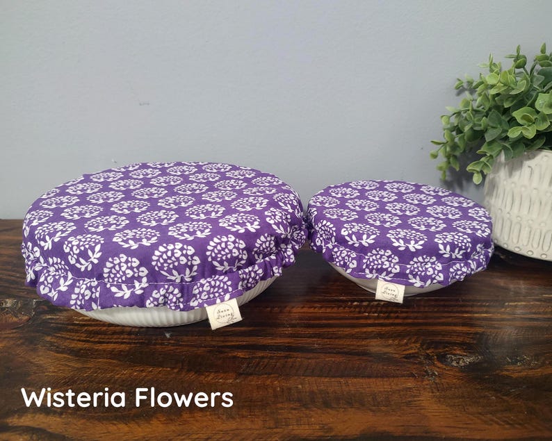 May include: Two round fabric bowl covers with an elasticized ruffled edge, featuring a purple and white floral pattern. Each cover has a small rectangular tag attached to the side with the text "Sara Living". The covers are placed over bowls.
