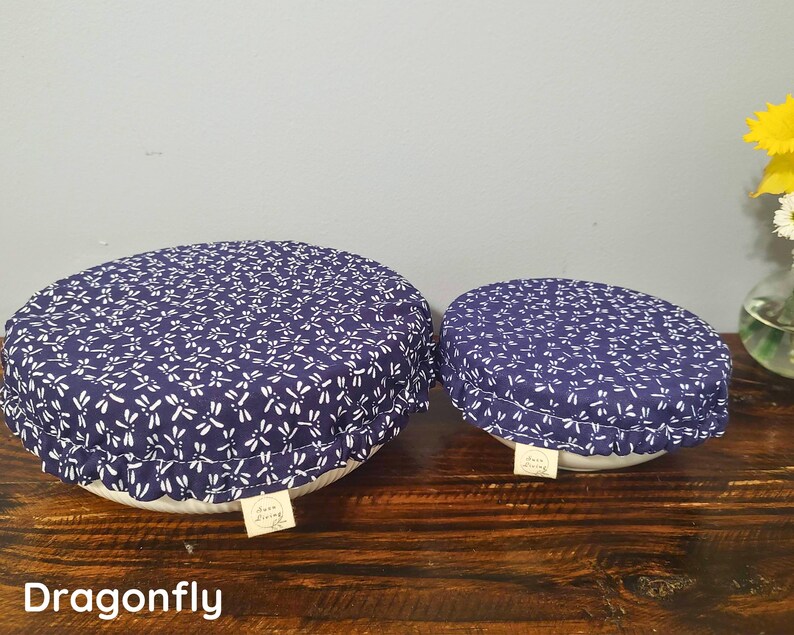 May include: Two round bowl covers made of dark blue fabric with a white dragonfly pattern. Each cover features an elasticized edge for a snug fit and a small rectangular beige fabric tag with the text "Aura Living" attached to the side.