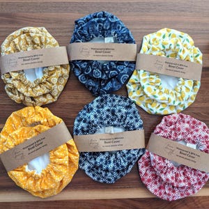 May include: Six reusable bowl covers with elastic edges, each wrapped in a brown paper band labeled Waterproof & BPA free Bowl Cover no more plastic wrap. The covers feature various patterns including yellow flowers, blue floral, yellow lemons, yellow dots, blue geometric, and red geometric prints.