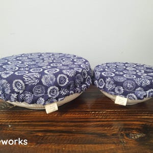May include: Two round fabric bowl covers with an elasticized edge, featuring a dark blue background with a white repeating floral or firework-like pattern. Each cover has a small white rectangular tag labeled Sara Living attached to the side.