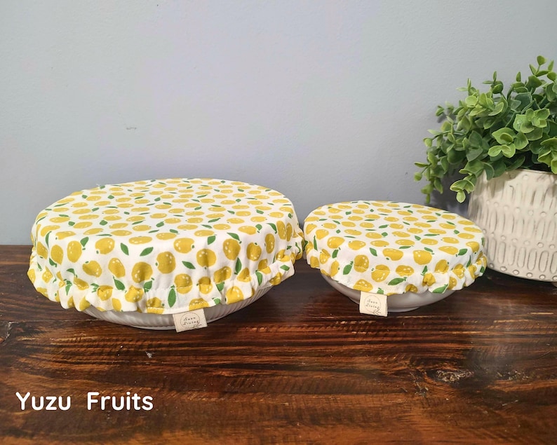 May include: Two round fabric bowl covers with an elasticized ruffled edge, featuring a white background with a repeating pattern of yellow yuzu fruits and green leaves. Each cover has a small rectangular fabric tag attached to the side.