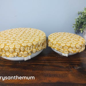 May include: Two round bowl covers with an elasticized edge, featuring a mustard yellow fabric with a white daisy-like floral pattern. Each cover has a small white rectangular tag attached to the side. The text Chrysanthemum is visible.