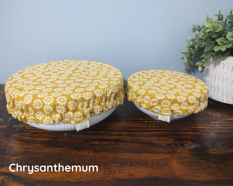 May include: Two round bowl covers with an elasticized edge, featuring a mustard yellow fabric with a white daisy-like floral pattern. Each cover has a small white rectangular tag attached to the side. The text Chrysanthemum is visible.