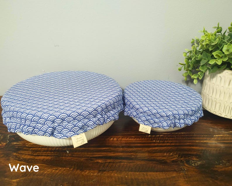 May include: Two round bowl covers featuring a blue and white wave pattern. Each cover has an elasticized edge and a small rectangular fabric tag. The covers are placed over white bowls, and the word Wave is printed in the bottom left corner.