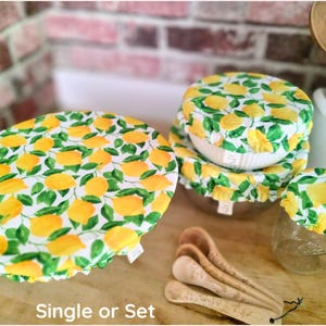 May include: A collection of fabric bowl covers featuring a yellow lemon and green leaf pattern on a white background. The set includes various sizes covering a large bowl, a smaller bowl, and a glass jar, with the text Single or Set visible.