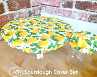 May include: A set of two fabric bowl covers featuring a yellow lemon and green leaf pattern on a white background. The larger cover is placed over a glass bowl, and the smaller cover is placed over a glass jar. A small white tag is visible.