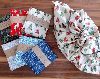 May include: A collection of fabric gift bags featuring various holiday patterns including Christmas trees, snowflakes, and stars. Five bags are folded flat with brown paper bands labeled Wrap for Life bag, while one larger bag is tied in a knot.