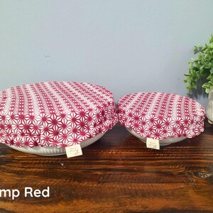 May include: Two round bowl covers featuring a red and white geometric star pattern. Each cover has an elasticized edge and a small white fabric tag with the text "Hemp Living" attached to the side. The covers are placed over white bowls.