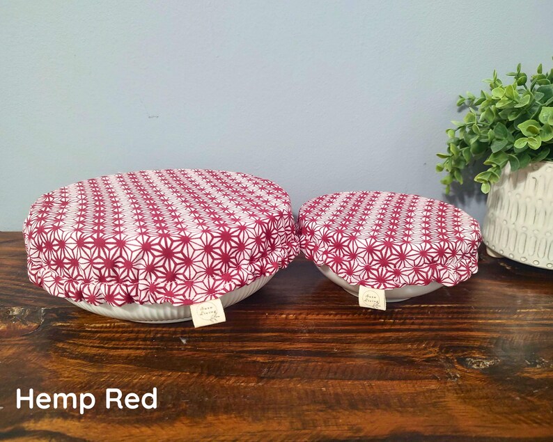 May include: Two round bowl covers featuring a red and white geometric star pattern. Each cover has an elasticized edge and a small white fabric tag with the text "Hemp Living" attached to the side. The covers are placed over white bowls.