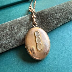 May include: A gold-toned oval locket pendant with a chain. The locket features a raised design of three interlocking circles. The chain and locket are displayed on a teal-colored surface.
