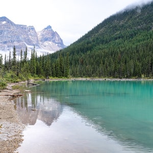 May include: A turquoise lake surrounded by a lush forest with snow-capped mountains in the background.