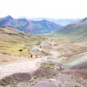 May include: A view of a valley in the Andes Mountains, with colorful rock formations and a dirt road leading down to a group of people.