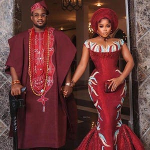 May include: A couple in formal attire, holding hands. The man wears a burgundy outfit with intricate embroidery and a matching hat, complemented by beaded necklaces. The woman is in a burgundy mermaid gown with blue and white accents, and a matching head wrap.