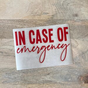 May include: A rectangular adhesive decal featuring the text IN CASE OF in bold red block capital letters, with the word emergency written below in a red cursive script font. The decal is presented on a translucent backing sheet.