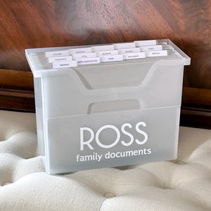 May include: A translucent plastic file box with white text reading ROSS family documents on the front. The box contains multiple hanging file folders with white tabs labelled with categories including banking, passports, utilities, vehicles, pets, birth certificates, receipts, home, school, insurance, taxes, and medical.