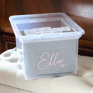 Peut inclure: Un bac de rangement en plastique transparent contenant des dossiers suspendus gris avec des onglets blancs étiquetés par niveau scolaire. L'avant du bac porte le prénom Ellie en lettres cursives rose clair, accompagné d'un petit cœur rose.