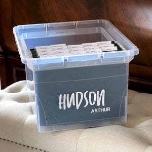 May include: A clear plastic storage bin containing dark blue hanging file folders. The front of the bin features the white text "HUDSON" and "ARTHUR". Inside, white file tabs are labelled with various grades and activities for organisation.