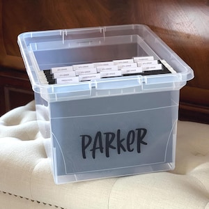 May include: A clear plastic storage bin containing black hanging file folders with white labels. The labels are printed with school grades from Preschool to 12th Grade and Yearbooks. The front of the bin features the name Parker in black text.