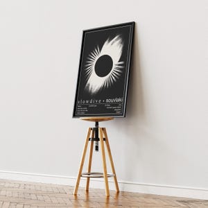May include: A framed black and white poster with a circular design and the text "slowdive &bull; souvlaki." The poster is displayed on a wooden stool with a black seat. The background is a white wall and a wooden floor. The stool is approximately 76 cm tall.