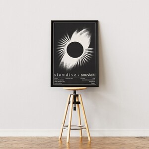 May include: A framed black and white poster of Slowdive's 'Souvlaki' album art. The design features a central black circle with radiating white lines. The poster is displayed on a wooden stool with a circular seat.