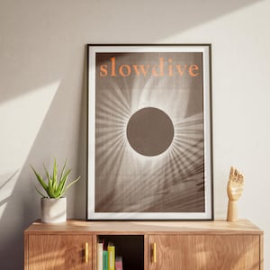Puede incluir: Impresi&oacute;n art&iacute;stica enmarcada con la palabra "slowdive" en naranja. La obra presenta un c&iacute;rculo oscuro con l&iacute;neas radiantes, similar a un eclipse solar. La impresi&oacute;n se exhibe en un mueble de madera con una planta y una escultura de mano de madera.