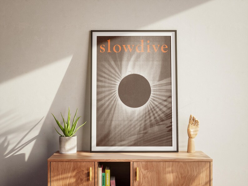 Puede incluir: Impresi&oacute;n art&iacute;stica enmarcada con la palabra "slowdive" en naranja. La obra presenta un c&iacute;rculo oscuro con l&iacute;neas radiantes, similar a un eclipse solar. La impresi&oacute;n se exhibe en un mueble de madera con una planta y una escultura de mano de madera.