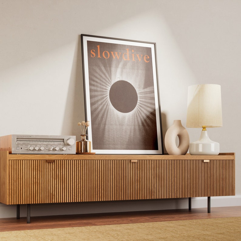 Puede incluir: Un mueble de TV de madera con frente acanalado, con un p&oacute;ster enmarcado con la palabra "slowdive" y un dise&ntilde;o circular. Una radio est&eacute;reo vintage, un jarr&oacute;n, una l&aacute;mpara y un jarr&oacute;n decorativo est&aacute;n encima. El mueble est&aacute; sobre patas negras.