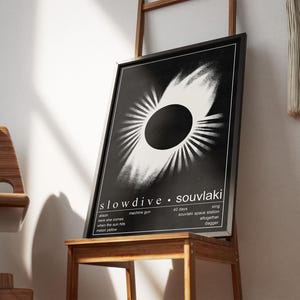 May include: A black framed art print with a white and black abstract design. The print, titled "slowdive souvlaki," is displayed on a wooden table. Song titles and lyrics are also printed on the artwork.