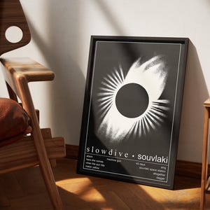 May include: A framed black and white art print with an abstract design, featuring a central circle and radiating lines. The print displays the text "slowdive &bull; souvlaki" and song titles. The artwork is positioned near a wooden chair.