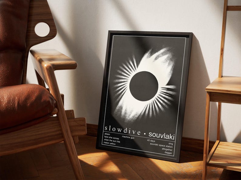 May include: A framed black and white art print with an abstract design, featuring a central circle and radiating lines. The print displays the text "slowdive &bull; souvlaki" and song titles. The artwork is positioned near a wooden chair.