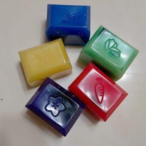 Puede incluir: Cinco jabones cuadrados de colores con diseños en relieve. Los jabones son azules, amarillos, verdes, rojos y morados. Cada jabón presenta un diseño diferente: una hoja, cerezas, una mariposa, una zanahoria y una flor.
