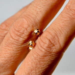 May include: A gold open ring featuring two small, faceted star-shaped ends, worn on a finger alongside a second ring that has a rectangular green gemstone surrounded by a border of small, clear, sparkling white stones set in a silver-toned band.