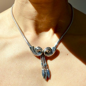 Op de afbeelding: Een zilveren ketting met een uniek hangerontwerp. De hanger heeft twee gebogen vormen die met elkaar verbonden zijn door een ketting met drie hangende traanvormige bedels.