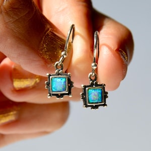 May include: A pair of drop earrings featuring square-cut iridescent blue opal stones set in silver frames. Each earring hangs from a simple silver hook, showcasing a vibrant play of colour within the square settings and a polished metal finish.