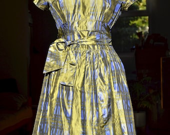Puede incluir: Vestido vintage sobre un maniquí. El vestido presenta una tela brillante con un patrón de azul y dorado. Tiene mangas cortas, un corpiño ajustado y una falda amplia. Una faja a juego está atada a la cintura, creando una silueta definida.