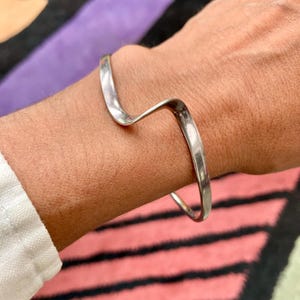 May include: A silver-colored, wave-shaped bracelet. The bracelet is worn on a wrist, with a simple, minimalist design. The metal has a polished finish, reflecting light. The background features a colorful, patterned rug.