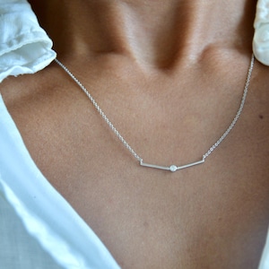 May include: A silver-toned necklace featuring a delicate chain and a horizontal bar pendant. The bar is slightly angled and holds a single, small, round, clear stone set in the center, creating a simple and refined piece of jewelry.