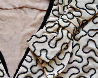 Chaqueta rara de los años 30, de color rosa beige, bien confeccionada, totalmente forrada, con bordados negros brillantes de estilo "garabato" y detalle de lazo. Talla pequeña.