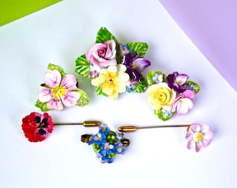 May include: A set of six vintage porcelain flower brooches. The brooches feature a variety of flowers, including roses, pansies, and forget-me-nots. The brooches are in shades of pink, yellow, purple, and blue.
