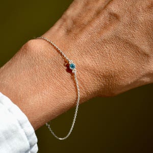 Könnte beinhalten: Ein zartes Silberkettenarmband mit einem einzelnen, kleinen, runden, in einer Zargenfassung gefassten blauen Edelstein in der Mitte. Das Armband besteht aus feinen, polierten Silbergliedern, die ein schlichtes und elegantes Erscheinungsbild bieten.
