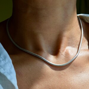 May include: A silver-toned snake chain necklace with a smooth, rounded profile and a flexible, interlocking link design. The metallic chain rests against the neck, featuring a consistent, polished finish throughout its entire length.