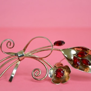 Könnte beinhalten: Eine goldfarbene Brosche in Form einer stilisierten Blume mit roten Glassteinen. Die Brosche hat ein filigranes, kunstvolles Design mit geschwungenen Linien und einer blattartigen Form.