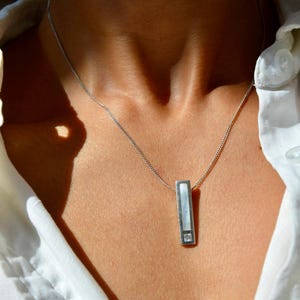 May include: Silver necklace with a rectangular pendant. The pendant has a white inlay and a small, square-cut gemstone at the bottom. The necklace is worn over a white collared shirt.